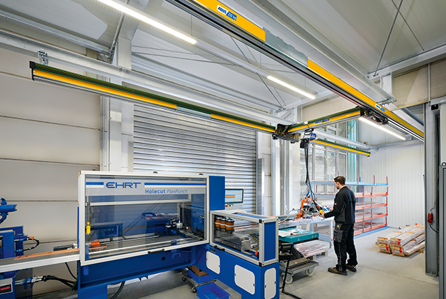 Le pont monopoutre EHB-X rehaussé déplace une charge dans un hangar industriel, tandis qu'un employé travaille sur une machine à l'arrière-plan.