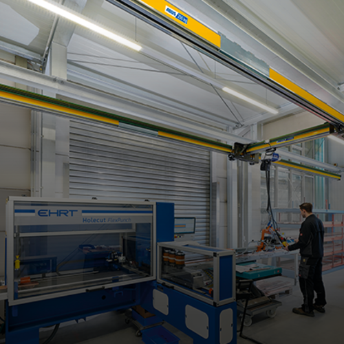 Le pont monopoutre EHB-X rehaussé déplace une charge dans un hangar industriel, tandis qu'un employé travaille sur une machine à l'arrière-plan.